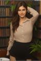 A woman in a beige shirt and black skirt posing in front of a bookshelf.